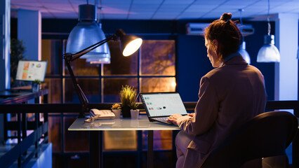 Caucasian woman planning business project at night working on computer with online analytics. Young employee sitting at desk analyzing e commerce statistics for report development. Handheld shot.