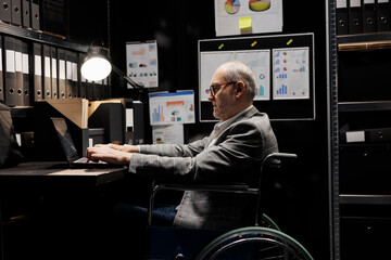 Senior businessman employee with disability in wheelchair inserting accountancy budget plan data on laptop. Bureaucratic executive in storage room office filled with chart reports and document folders - obrazy, fototapety, plakaty