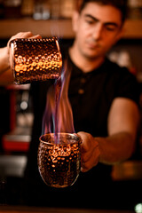Handsome male bartender pouring burning alcoholic drink from one steel cup to another