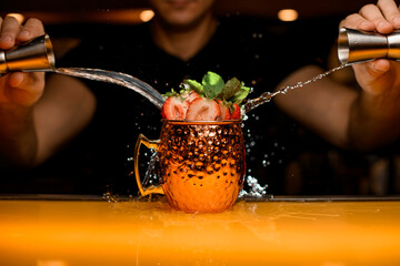Fototapeta Male bartender pours a jigger drink into cocktail cup decorated with strawberry and mint leaves obraz