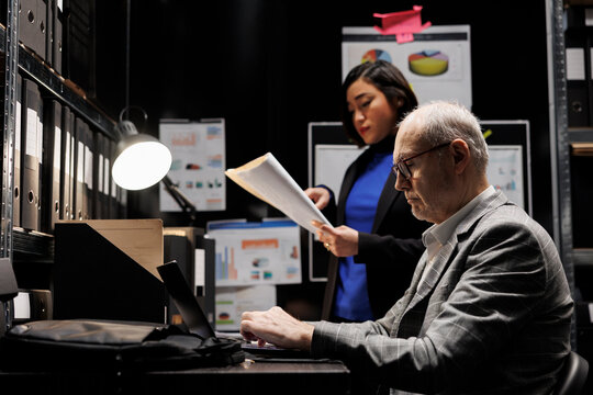 Professional businesswoman and coworker accountancy employees imputing bureaucratic market data paperwork on laptop. File cabinet room filled with document folders and flowcharts - Powered by Adobe