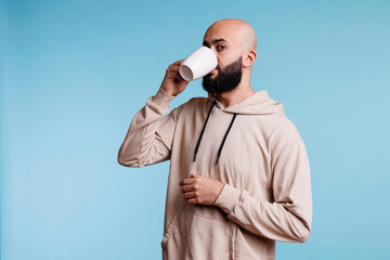 Young arab man drinking coffee from white cup and looking at camera. Arabian bald bearded person wearing casual clothes sipping tea, enjoying hot beverage from mug studio portrait