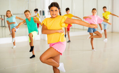 Portrait of cheerful preteen hispanic girl learning energetic dance moves with group of children in...