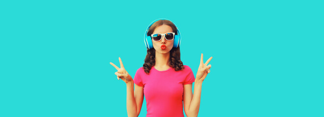 Portrait of cool teenager girl in headphones listening to music blowing her lips sending air kiss on blue background