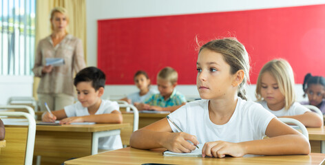 Naklejka na ściany i meble Portrait of positive small female pupil sitting at desk studying in classroom