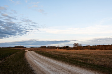 Winding dirt country road. Landscape of agricultural fields in spring. Evening blue sky with gray and white clouds. A twilight shadow falls to the ground