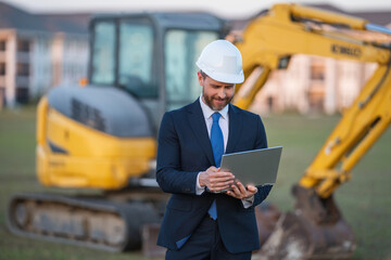 Construction owner near excavator. Confident construction owner in front of house. Architect, civil engineer. Man construction owner with a safety vest and hardhat at construction site.