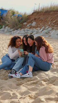 Four diverse young girls using phone together while sitting at beach