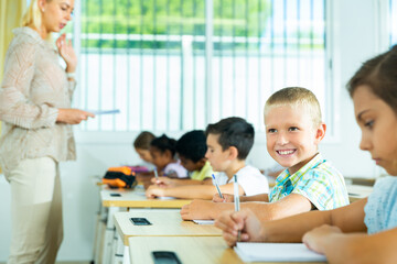 Portrait of cheerful cute blond tween schoolboy sitting at desk in classroom during lesson
