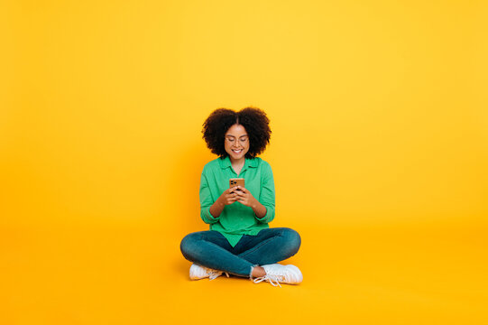 Full-length photo of happy positive trendy african american or brazilian curly woman, sits on a yellow background, uses her smartphone, chats on social networks, browsing internet, news, smiles