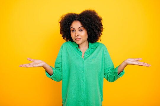 Confused hispanic or brazilian lovely curly haired woman, wearing green shirt, gesticulates with his hands to the side, puzzled, looks inquiringly at the camera, stand on isolated yellow background