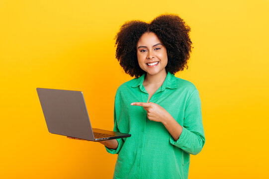 Friendly gorgeous brazilian or african american curly haired woman, in a green shirt, holding an open laptop in hand, points finger at it, looks at camera, smiles, stand on isolated orange background
