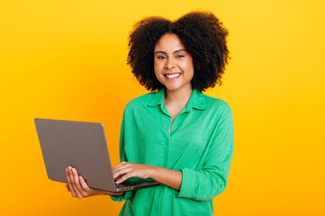 Positive beautiful brazilian or african american curly haired woman, dressed in a green shirt, holding an open laptop in hand, looking at camera, smiling, standing over isolated orange background