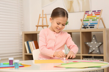 Cute little girl using glue stick at desk in room. Home workplace - obrazy, fototapety, plakaty