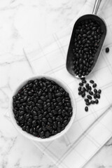 Bowl and scoop of raw black beans on white marble table, flat lay