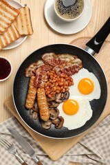 Frying pan with cooked traditional English breakfast and cup of coffee on wooden table, flat lay
