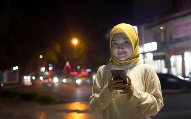 Beautiful Asian Moslem Young Woman Using Smartphone Walking Through Night City Street Full of Neon Light. Portrait of Gorgeous Smiling Female Using Mobile Phone.