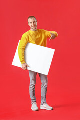 Young man pointing at big blank poster on red background