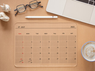 Top view, flat lay, the online calendar on a transparent tablet computer showing screen July 2023, on a brown background, coffee cup, laptop, and eyeglass. Reminder for schedule planning..