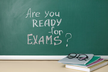 Notebooks and eyeglasses on table against blackboard with question ARE YOU READY FOR EXAMS?