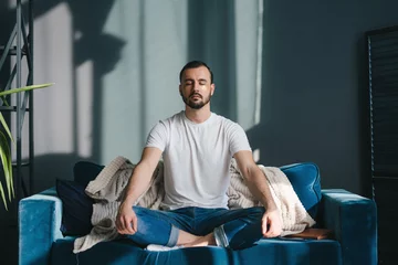 Tuinposter Focused man meditating in lotus pose breathing deep and slowly wearing casual clothes sitting on comfortable coach relaxing at home. Feeling free from stress. © Strelciuc