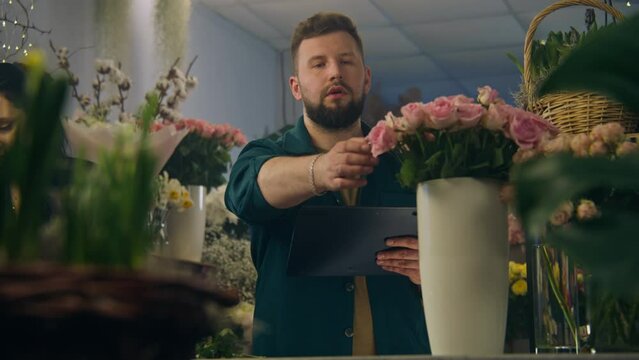 Male florist counts flowers in vase and takes order online in floral shop using digital tablet computer. Female colleague collects bouquet nearby. Flower small business and entrepreneurship concept.