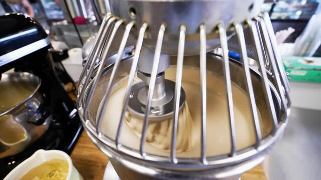 Stainless steel commercial stand mixer rapidly whisks batter, numerous stand mixers of various sizes clutter bakery kitchen counter, slow motion 4K