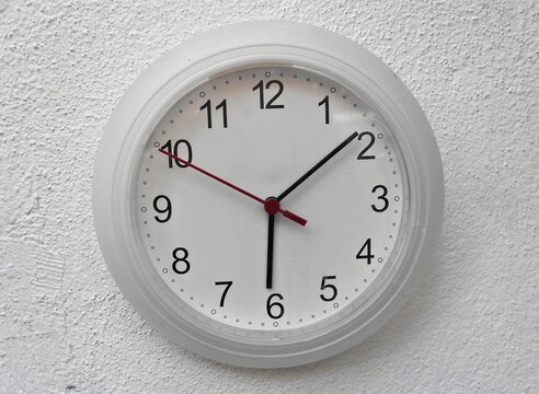 The picture displays a white wall clock, prominently showing the current time with its distinct hands and numerals, against a neutral background.