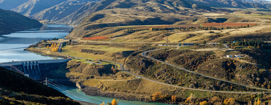 Panoramic aerial view to Clyde dam with river and winding road on hillside surrounded by mountainous and autumn trees.
