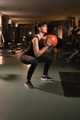 Fototapeta na wymiar Working out at gym. A young woman squats working out muscles by holding the medicine ball sitting in the modern gym. Health and fitness