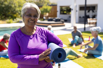 Portrait of smiling senior biracial woman holding yoga mat, with diverse yoga group in sunny garden - Powered by Adobe