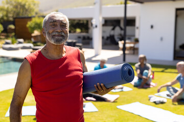 Portrait of smiling senior biracial man holding yoga mat, with diverse yoga group in sunny garden