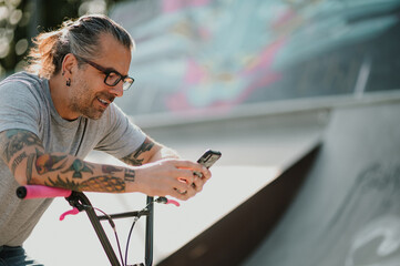 A tattooed middle-aged urban man is sitting on his bmx bike in a skate park and watching at videos on social media.