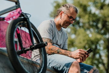 A happy mature tattooed urban man is sitting in a skate park next to his bmx bike and smiling at...