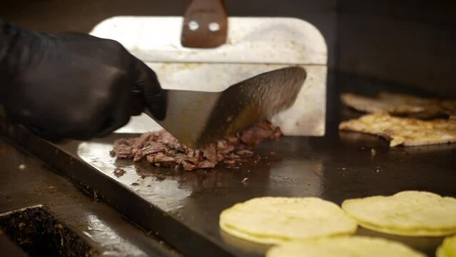 Cook cuts through chopped beef fajita meat with edge of spatula, flips taco meat on flat top griddle as corn tortillas heat up, slow motion close up 4K