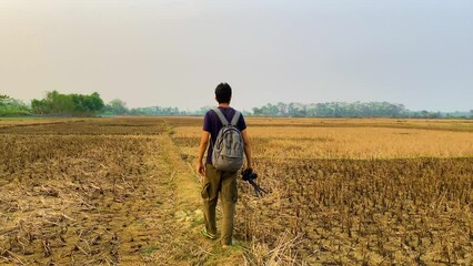Slow Motion Shot of a Photographer Walking on a Path Along Dry Fields, Holding a DSLR and Wearing a Backpack, With Trees in the Background (UHD)