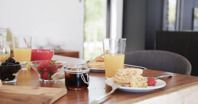 Breakfast pancakes on plates, with honey, fruit and juice on table in sunny kitchen, slow motion