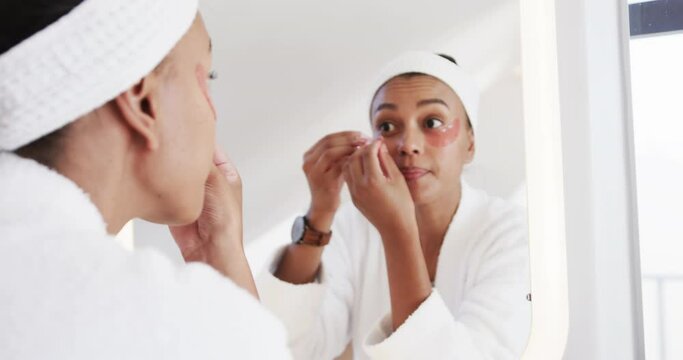 Focused biracial woman applying under eye masks looking in mirror in sunny bathroom, slow motion