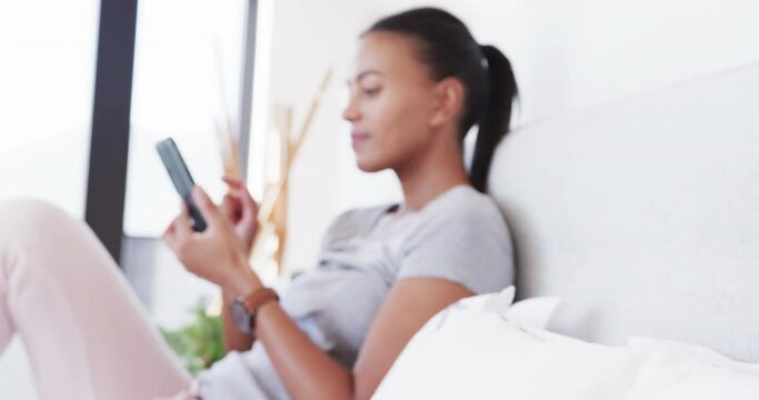 Happy biracial woman sitting on bed using smartphone, in slow motion