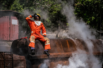 Professional firefighter is sitting on a rusty building holding a water glass and drinking at an outdoor site among the smoke.