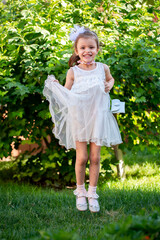 A cheerful little girl plays in the park on the lawn in a beautiful dress rejoices in the summer. Beautiful white girl smiling while walking in the courtyard of the house.