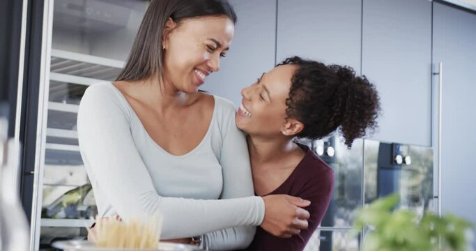 Happy biracial lesbian couple preparing food, embracing and laughing in kitchen, in slow motion