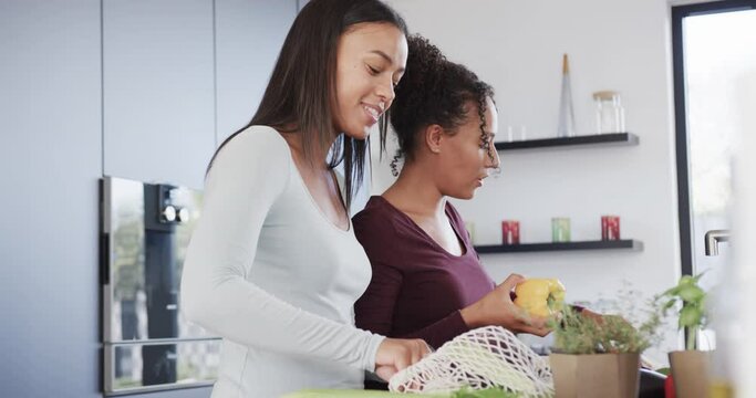 Happy biracial lesbian couple unpacking grocery shopping and talking in kitchen, in slow motion