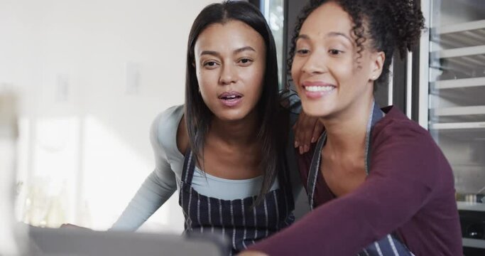Happy biracial lesbian couple in aprons preparing food and using tablet in kitchen, in slow motion