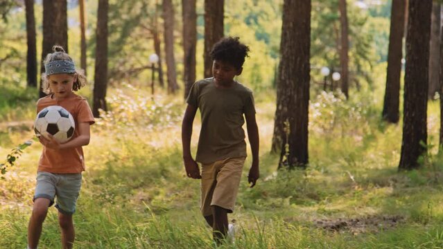 Full shot of African American and Caucasian elementary age boys walking in woods with ball in hands and talking