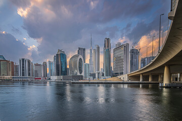 Obraz premium Sunset with a cloudy sky over the financial district of Dubai. City skyline in business center with office buildings and skyscrapers around Burj Khalifa. Street with a bridge over the harbor