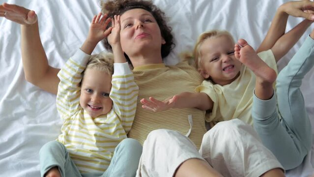 Top view slow motion happy cheerful woman with children on bed dangling their legs smiling laughing playing. Happy weekend day off with family. Woman and children on bed dangle their legs having fun