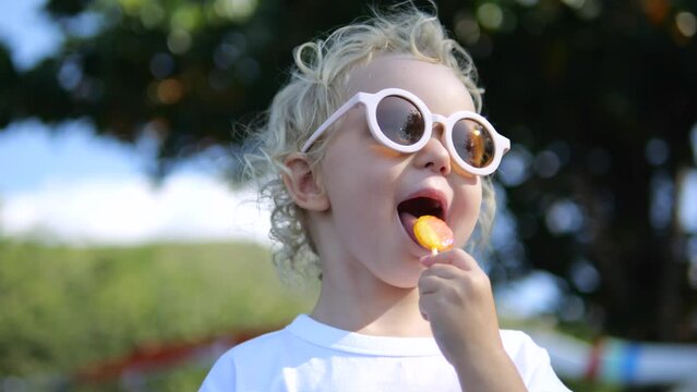 Low angle shot, Caucasian child in cool fashionable sunglasses licks sugary sweet lollipop, sweetness on hot summer day. Child licks lollipop while standing in park in sun, low angle close-up shot.