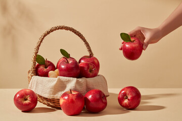 Red ripe apples are placed in a bamboo basket, some fall on a beige background. The female hand holding a small apple with green leaf that is being added to the basket.