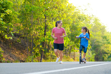 Active little girl riding a scooter on a park road on a summer day with her mother. Outdoor sports for children. Family spending time together